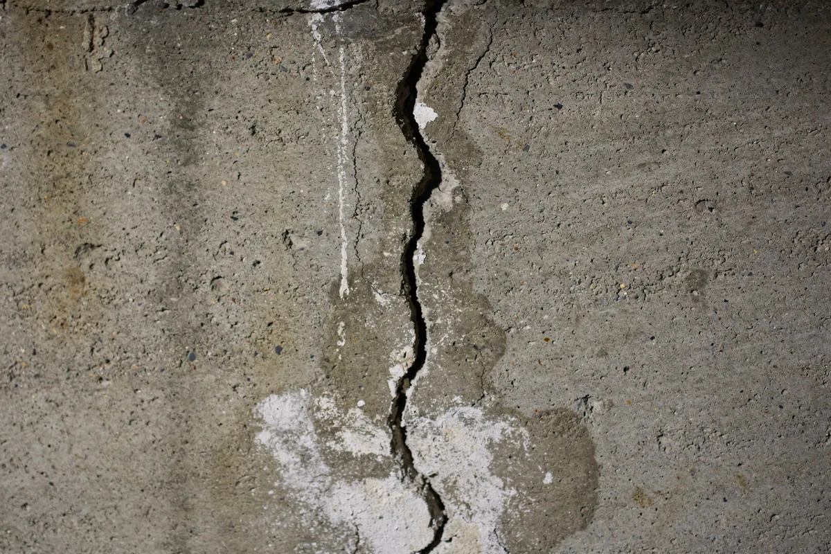 A narrow vertical crack in a poured concrete basement wall with a faint damp stain and pale mineral deposit trailing down from it.