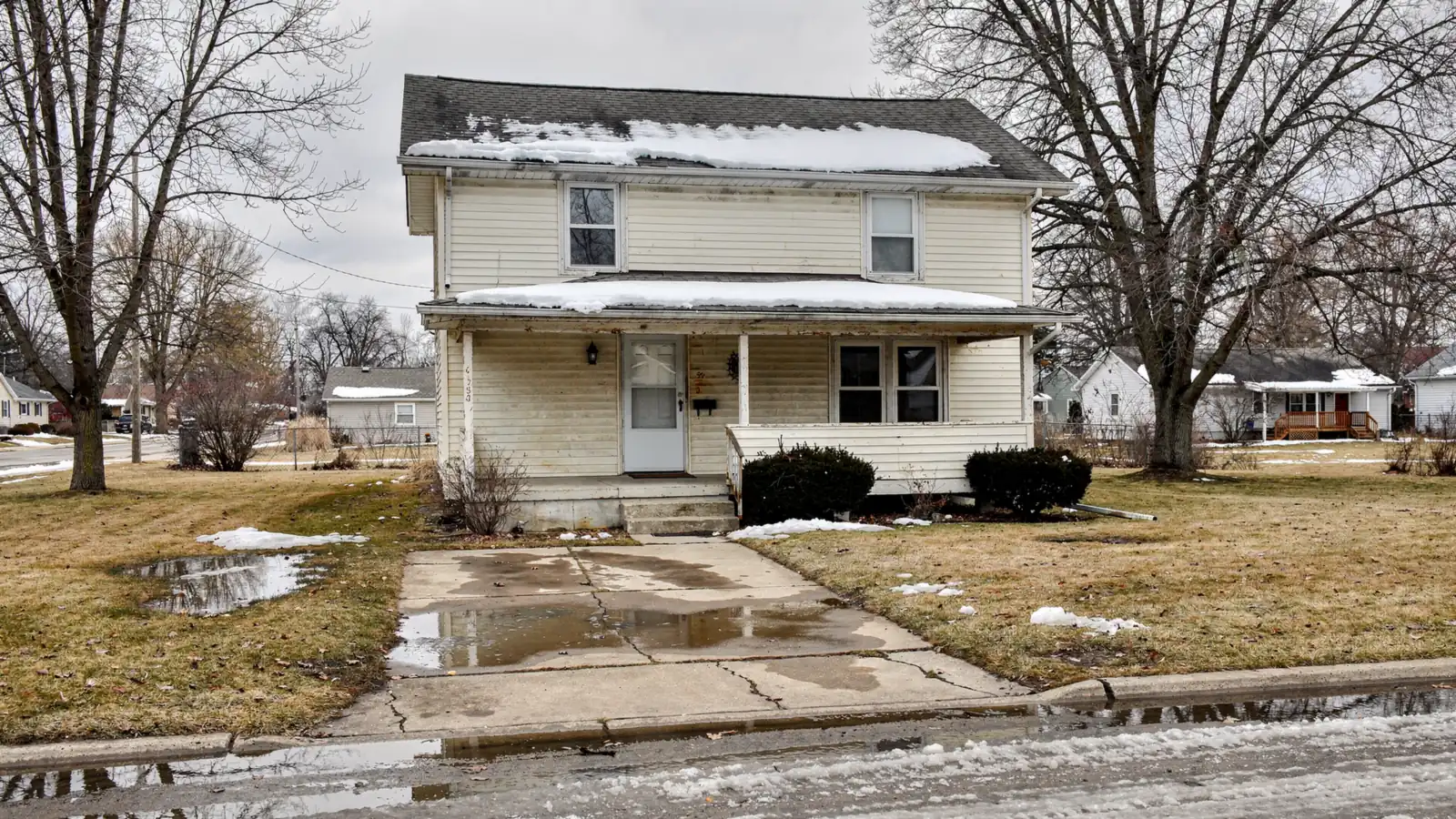 A plain two storey clapboard house on a flat lot with snowmelt standing across the lawn and driveway under a grey late winter sky.